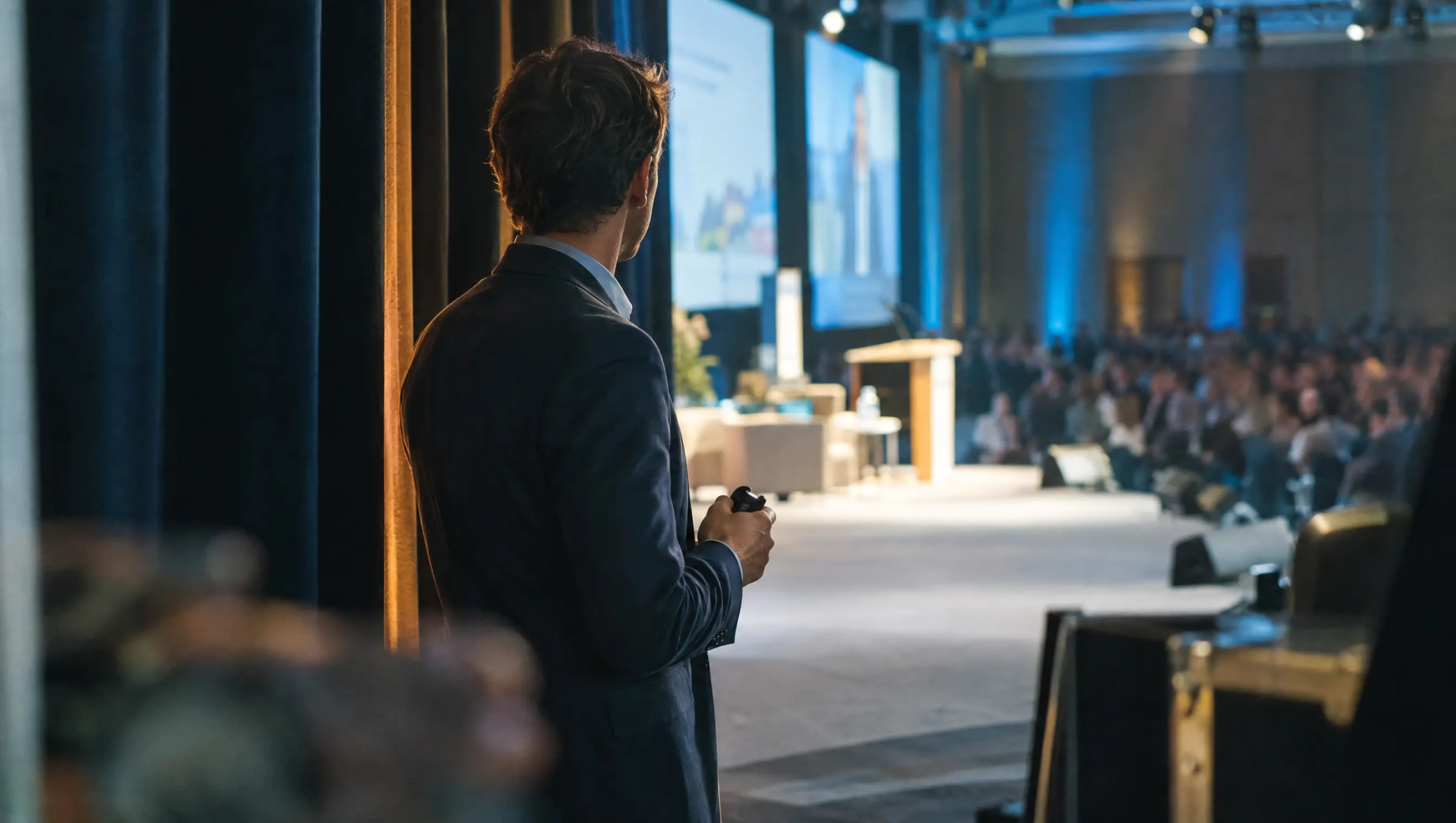 A speaker waiting in the wings of a conference stage, seen from behind