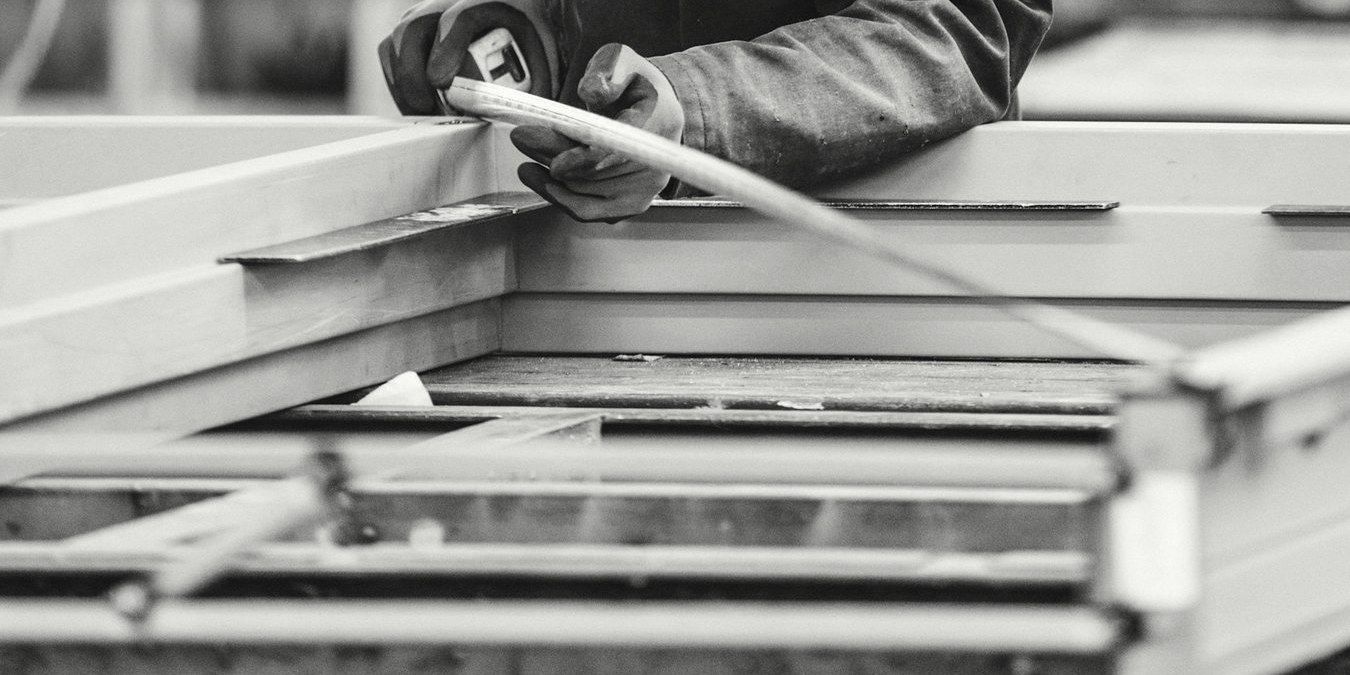 Gloved hands measuring an aluminium frame on a workshop bench